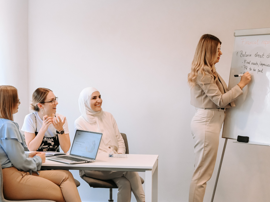 A group of women collaborate in a meeting as one presents financial notes on a whiteboard.