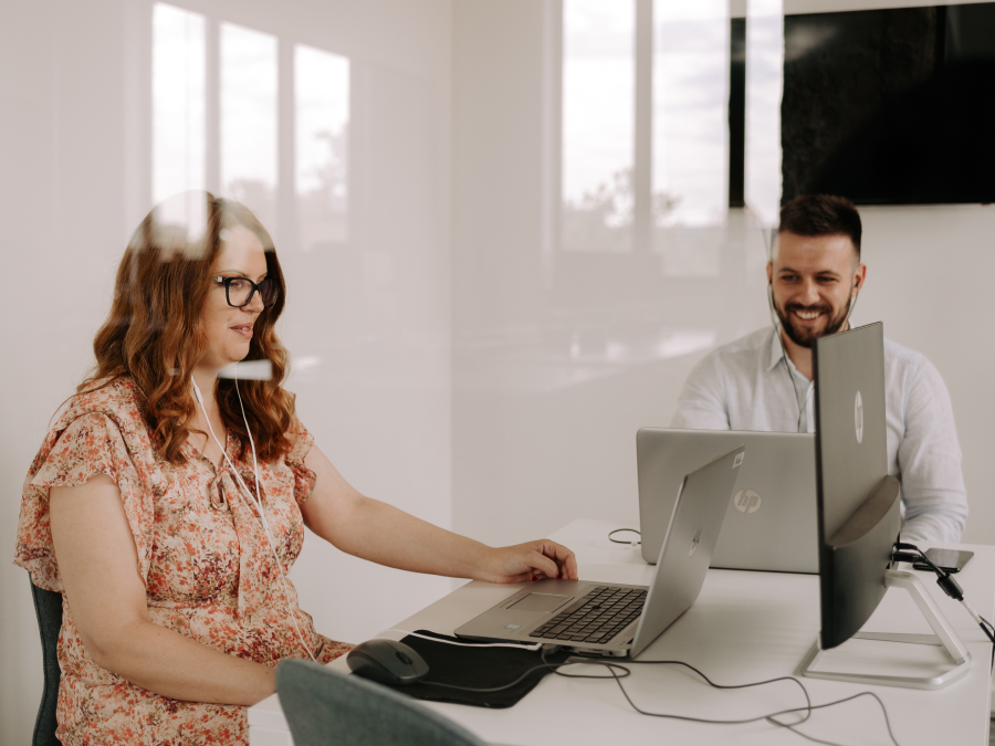 A team leader sits with a teammate in a meeting room, assisting her with a task on her laptop.