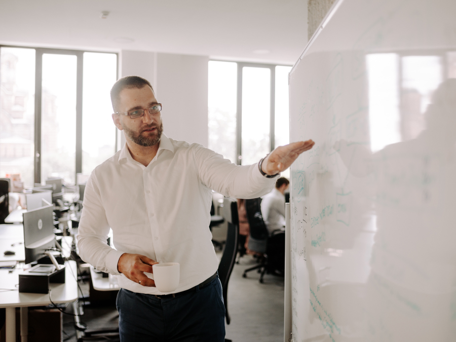 The image shows a man in an office explaining ideas on a whiteboard while holding a cup, representing planning, organization, and effective time management skills.